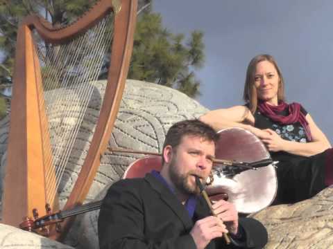 Neil playing tin whistle and Christina beside her cello with Nei;'s Harp behind all sitting on rocks outdoors
